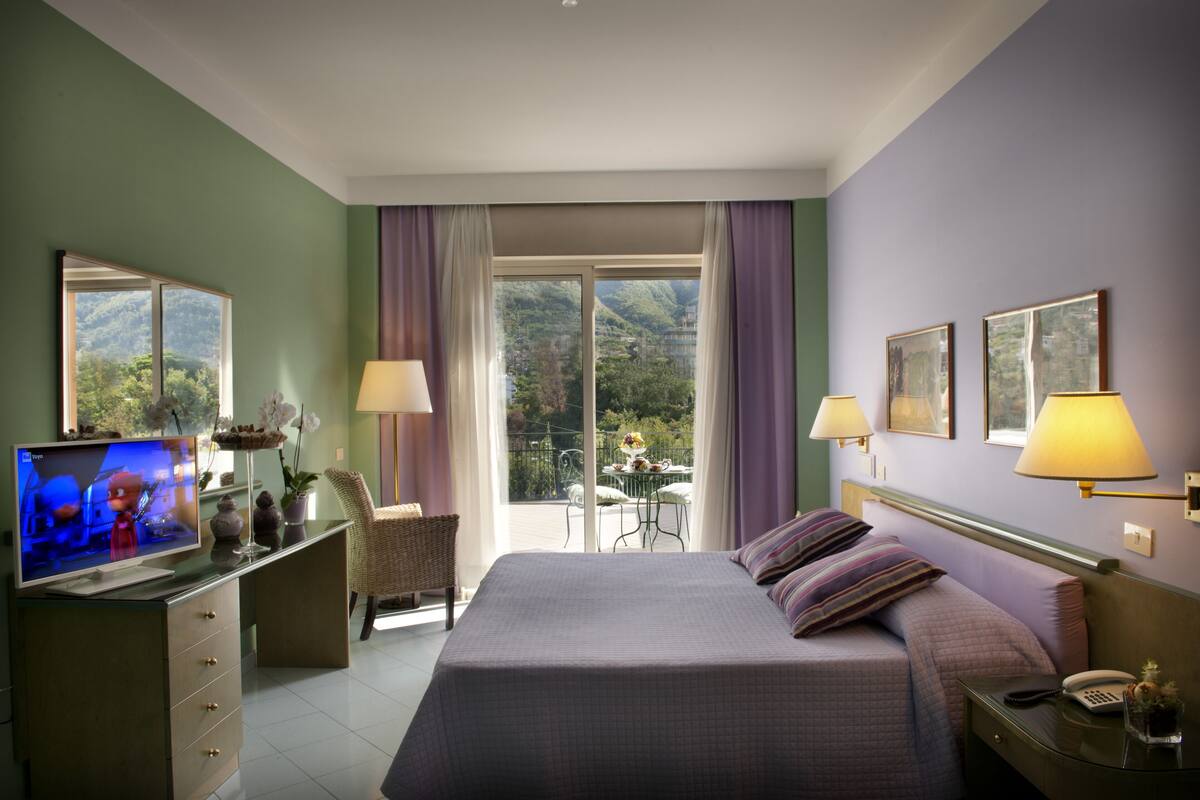 Een hotelkamer met een bed, een televisie, een bureau met een computer, een stoel, een tafel met een vaas, en een balkon met uitzicht op de bergen.
