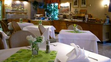 A dining area with tables set for service, a kitchen in the background, and a view of the outdoors.