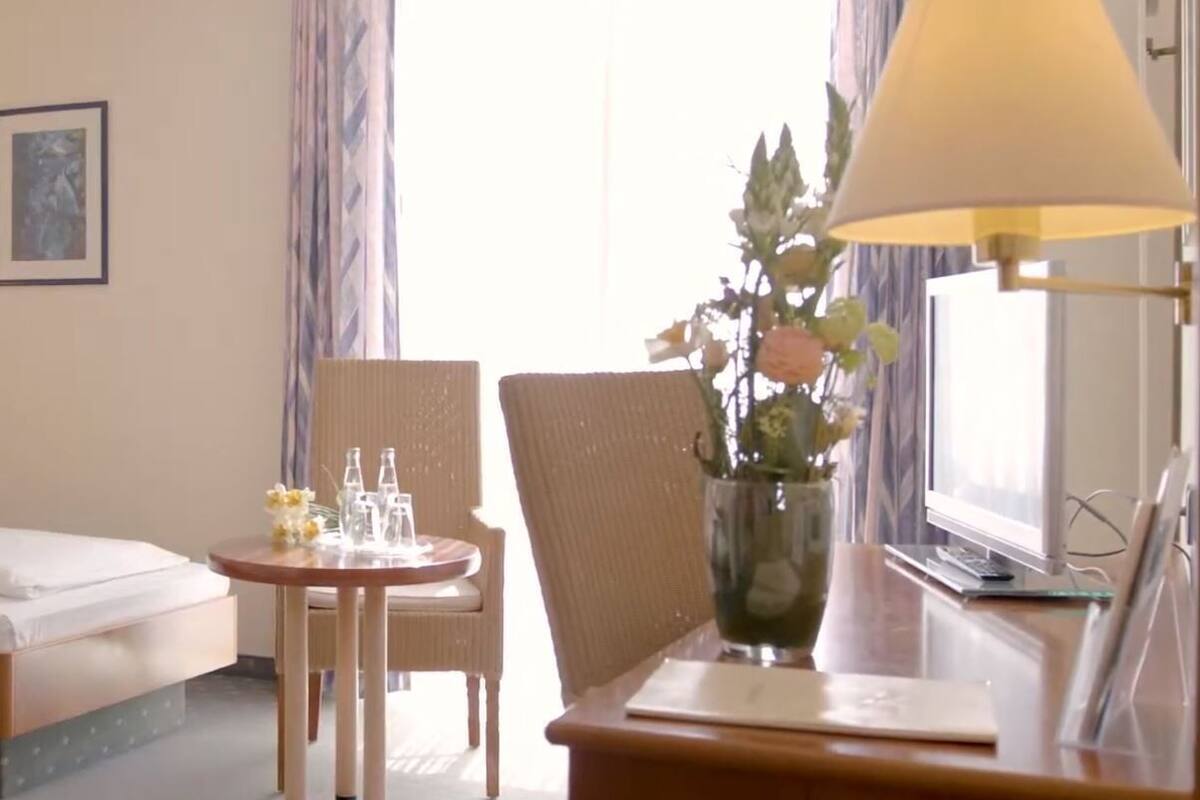 Ein Hotelzimmer mit zwei Betten, einem kleinen Tisch mit Blumenvase, einem Sessel und einem Schreibtisch mit Lampe.