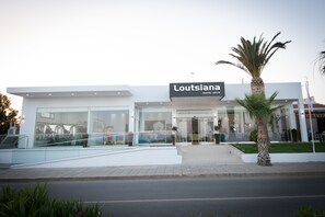 A modern hotel named 'Loutisiana' with a glass facade, a palm tree, and a well-maintained sidewalk. - Loutsiana Hotel Apts (Ayia Napa)
