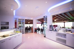 A modern cafe with a clean, white interior, a coffee machine, and a seating area with tables and chairs. - Loutsiana Hotel Apts (Ayia Napa)