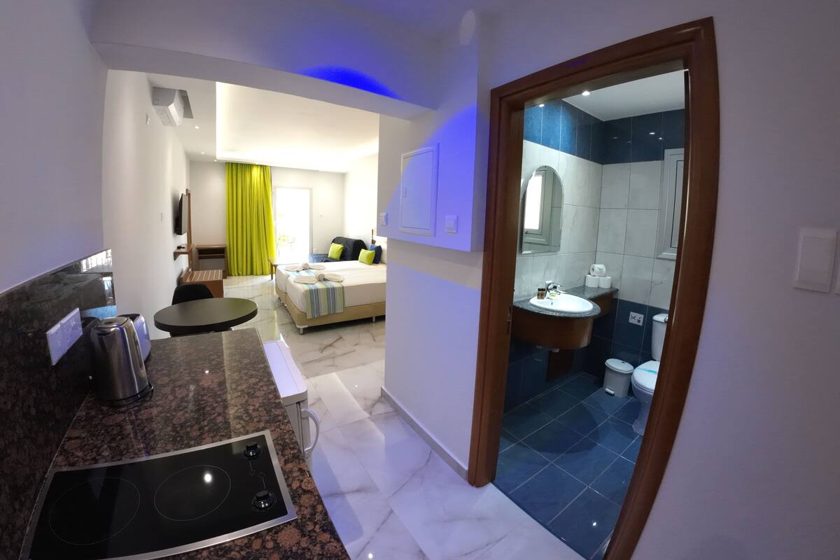 A modern hotel room with a kitchen area, a dining table, a bed with pillows, and a bathroom with a sink and toilet.