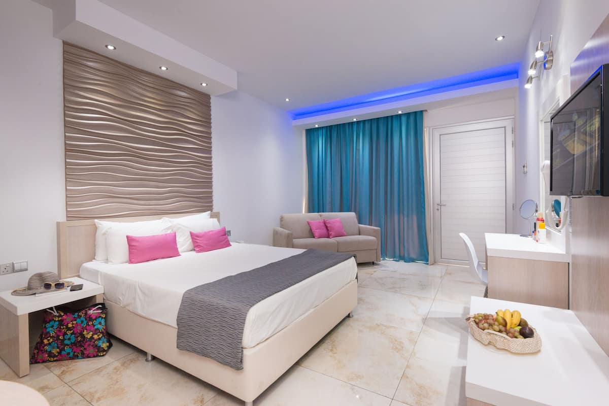 A modern hotel room with a large bed, a sofa, a desk with a fruit bowl, a TV, and a bathroom with a shower curtain.