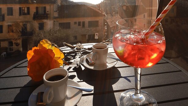 A table with a cup of coffee, a glass of red drink, and a flower on a balcony overlooking a town with buildings and mountains in the background. - Hotel Napoléon (La Turbie)