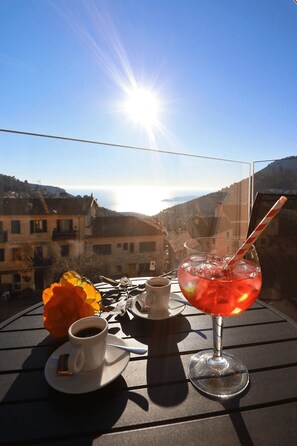 Una mesa con una taza de café, un vaso de bebida roja y una flor en un balcón con vistas a un pueblo y montañas al fondo.