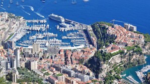 An aerial view of a coastal city with a marina filled with boats, historic buildings, and a prominent castle-like structure. - Hotel Napoléon (La Turbie)