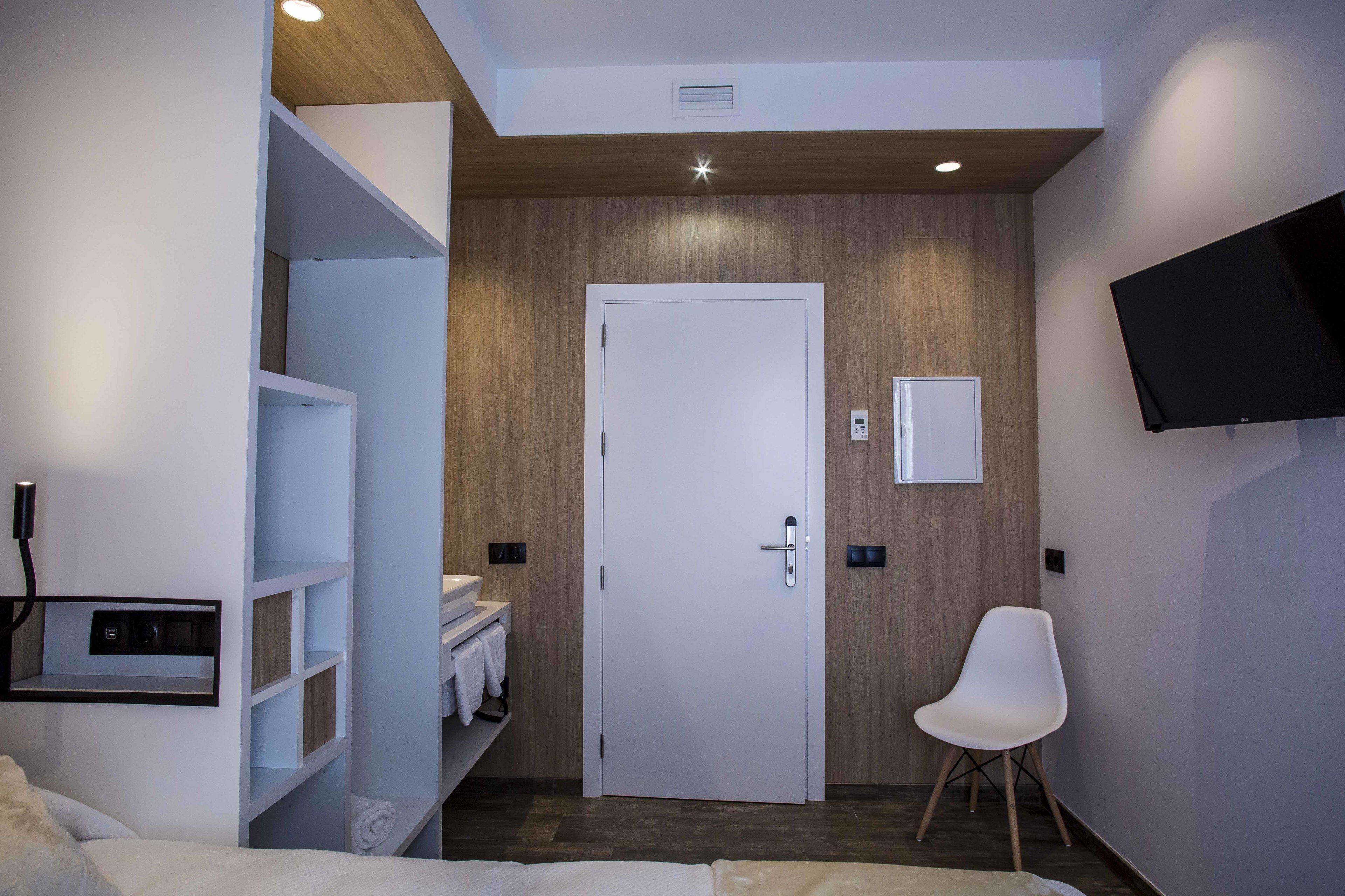 A modern hotel room with a wooden accent wall, a white door, a flat-screen TV mounted on the wall, a white chair, and a built-in shelf with towels.