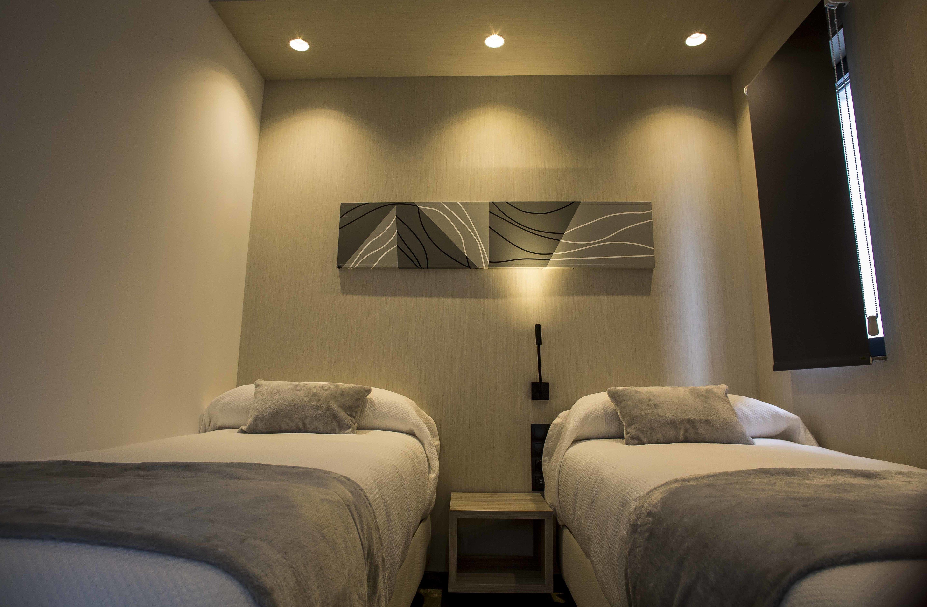 A hotel room with two beds, a wall-mounted artwork, a bedside table, and a window with blinds.