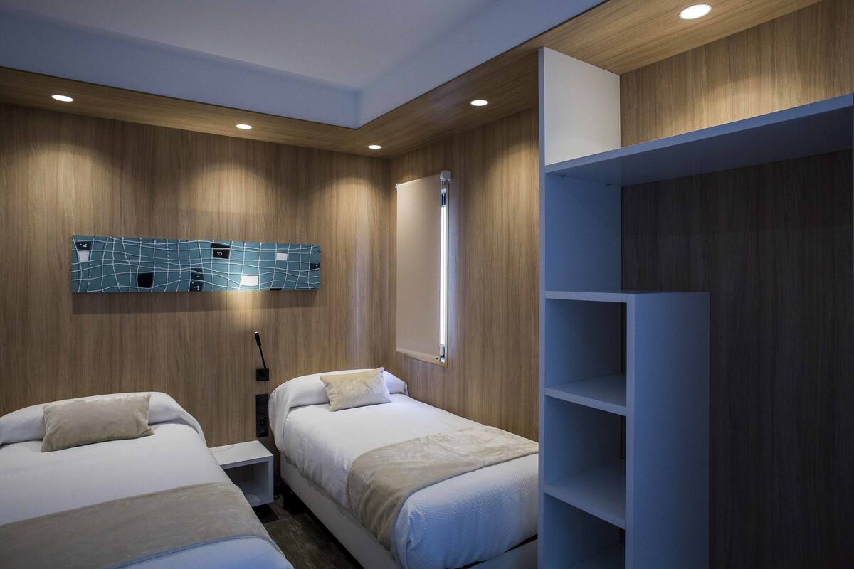 A modern hotel room with two beds, a wooden wall panel, a blue geometric wall art, a white wardrobe, and a bedside table with a lamp.