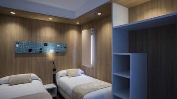 A modern hotel room with two beds, a wooden wall panel, a blue geometric wall art, a white wardrobe, and a bedside table with a lamp.