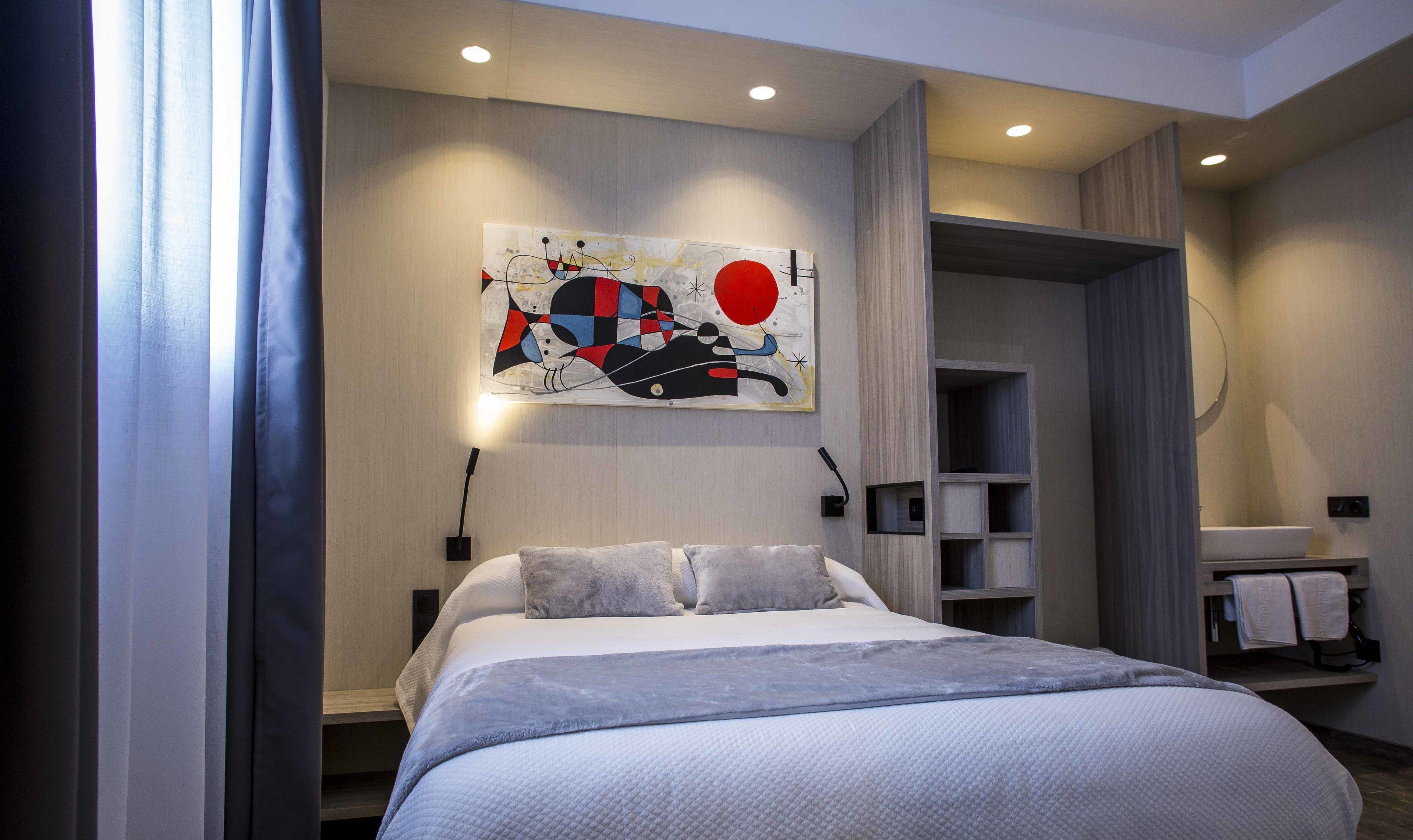 A modern hotel room with a large bed, a wall-mounted abstract painting, built-in shelving, and a bathroom with a round mirror.