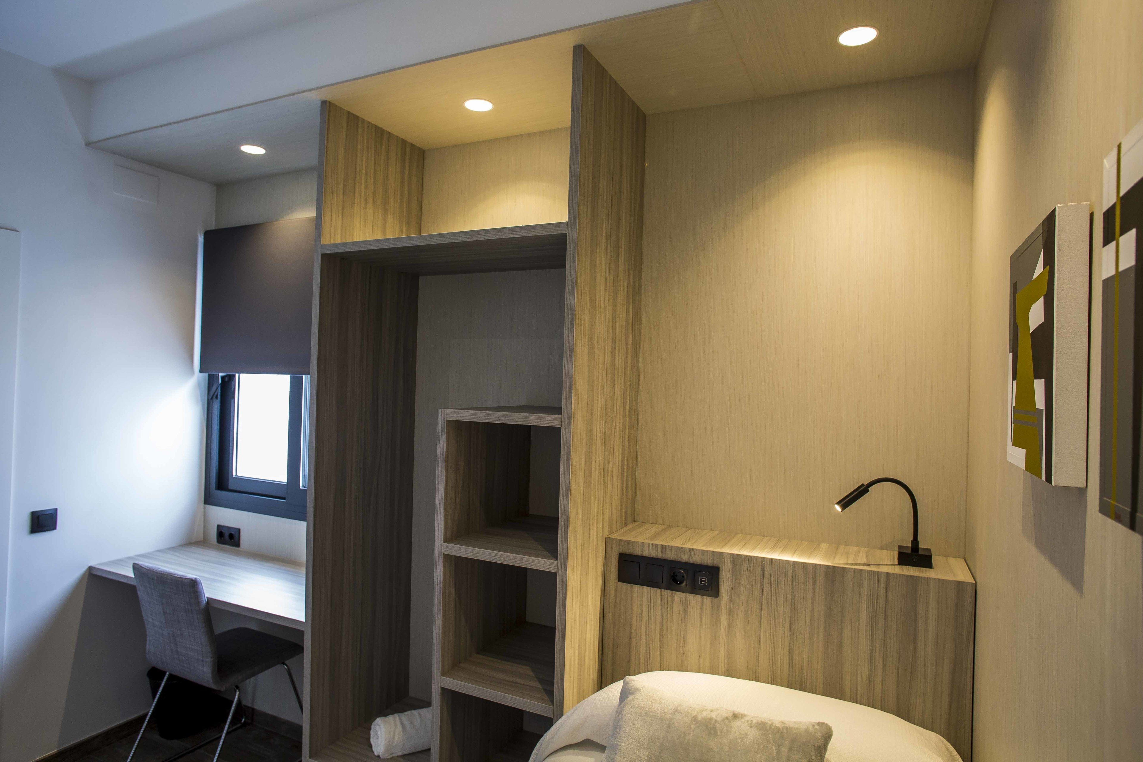 A modern hotel room with a desk, a chair, a bed, and built-in shelving.