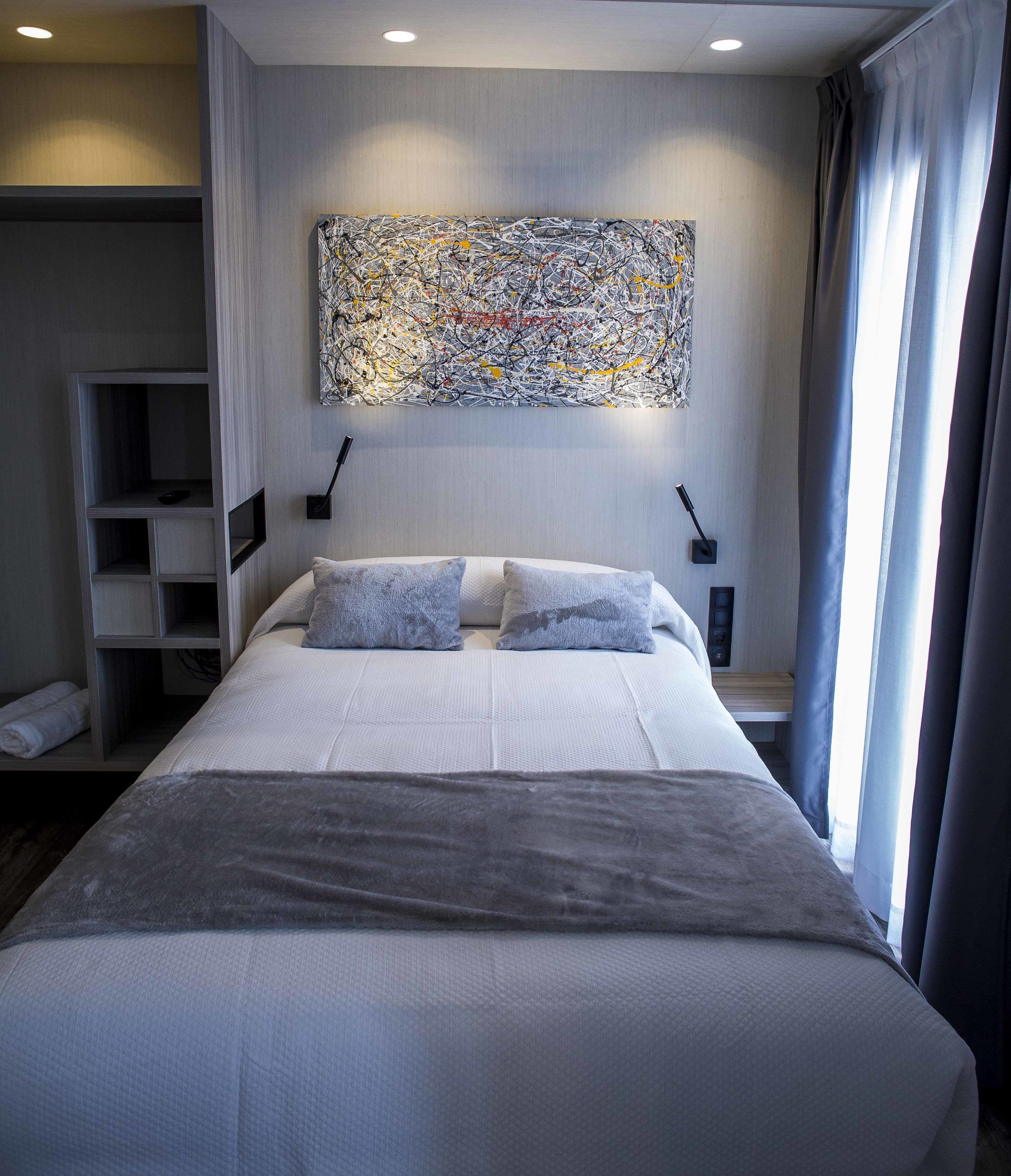 Una habitación de hotel moderna con una cama grande, una mesita de noche, un velador, un cuadro abstracto colgado en la pared y una ventana con cortinas.