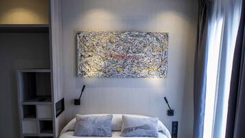 A modern hotel room with a large bed, a bedside table, a nightstand, a wall-mounted abstract painting, and a window with curtains.