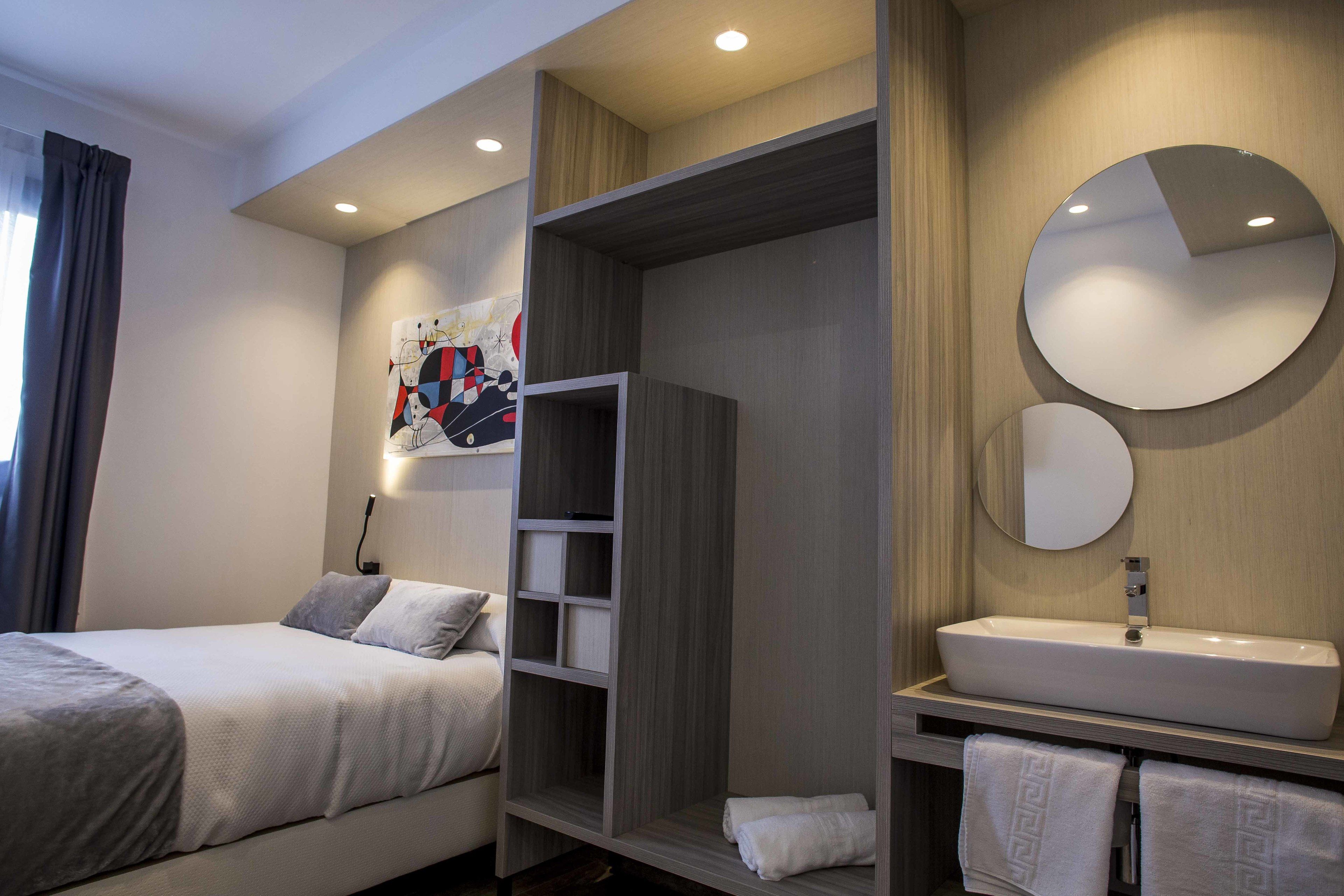 Una habitación de hotel moderna con una cama, un armario, un baño con lavamanos y espejo, y un cuadro en la pared.