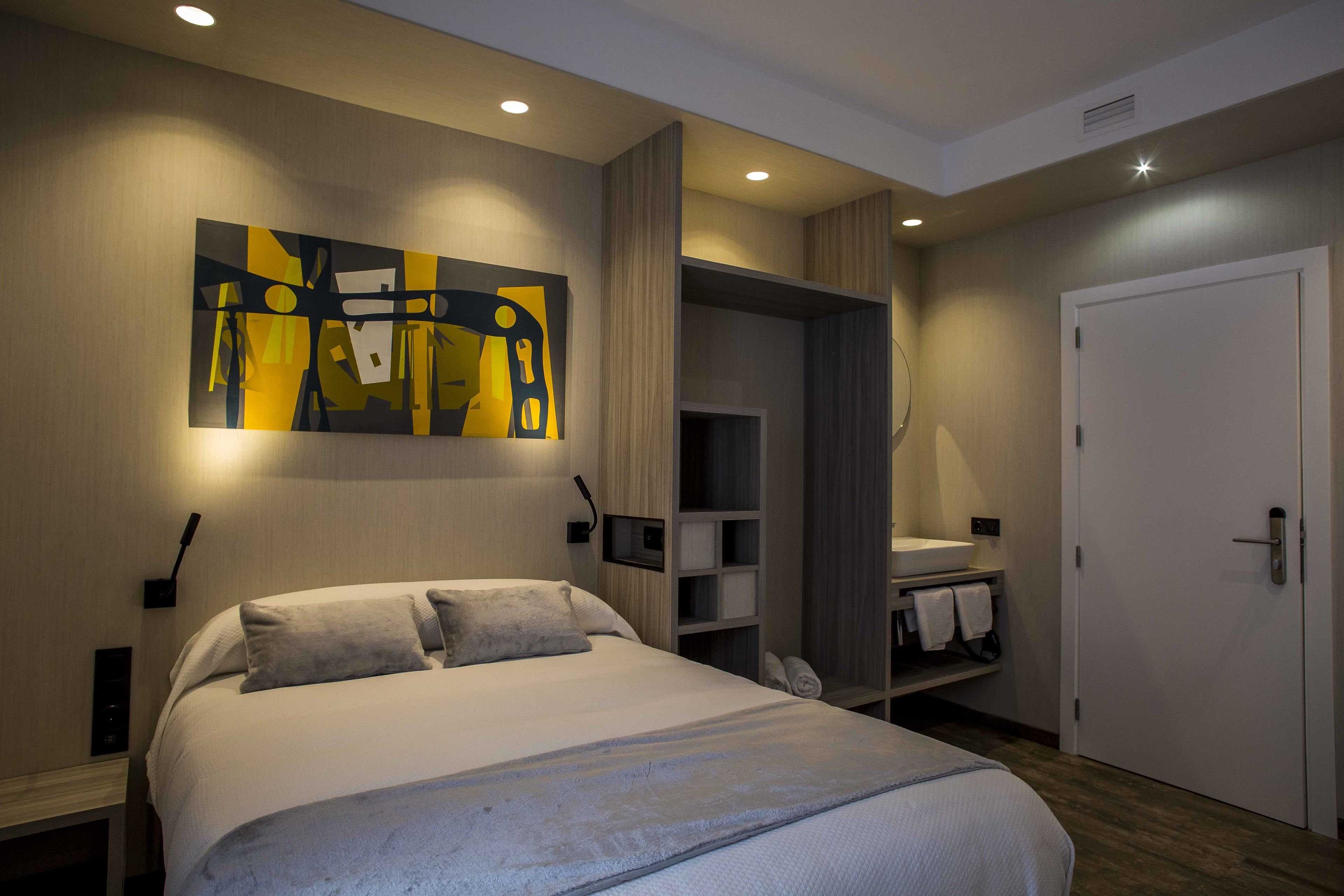 A modern hotel room with a large bed, a painting on the wall, a walk-in closet, and a bathroom with a sink and towel rack.