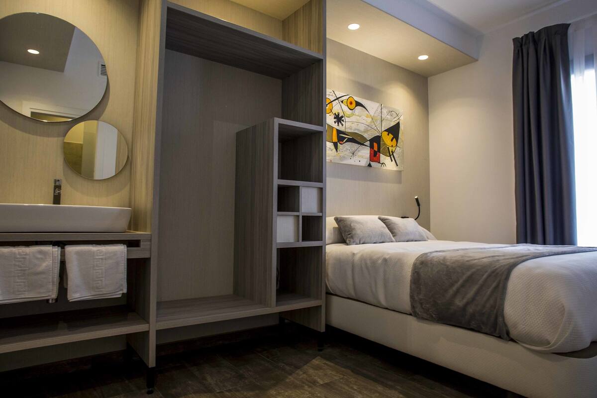 A modern hotel room with a bed, a built-in wardrobe, a bathroom with a sink and mirror, and a wall art piece.