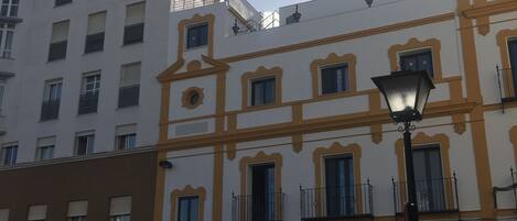 A street view with a building featuring a white facade with yellow trim and multiple windows. There is a street lamp in the foreground.
