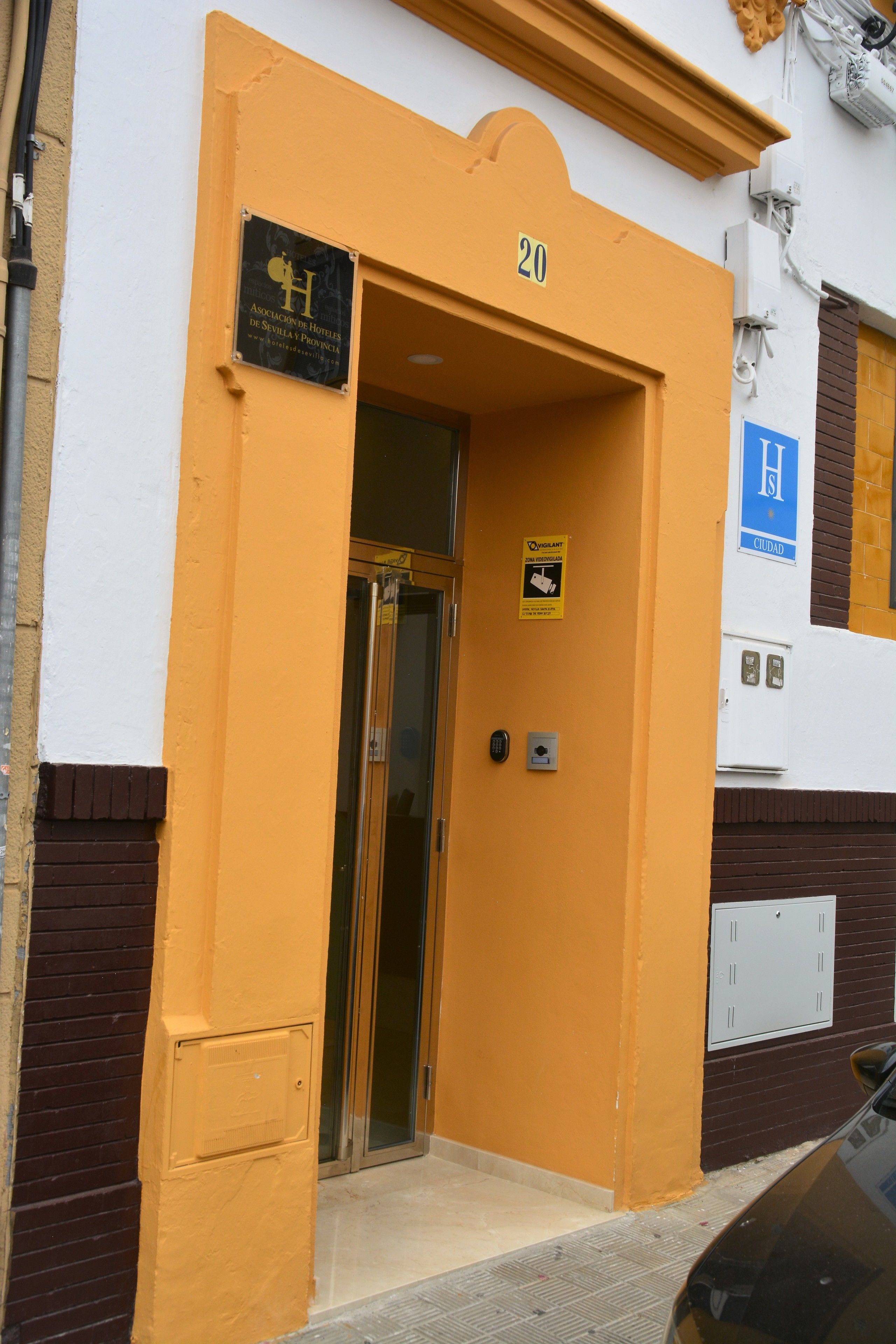A hotel entrance with a yellow facade, a door with a glass panel, and a sign indicating the hotel's name and room number.