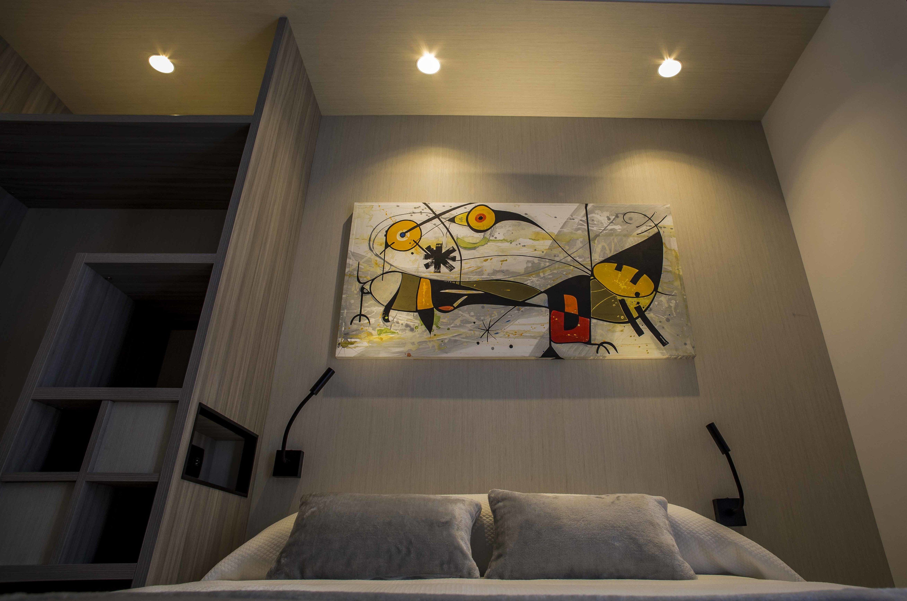 Et moderne hotelværelse med et stort abstrakt billede på væggen, en seng med gråt sengetøj og indbyggede hylder.