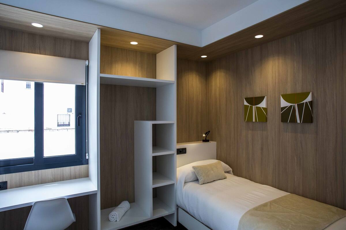 A modern hotel room with a bed, a desk with built-in shelving, a window, and two abstract wall art pieces.