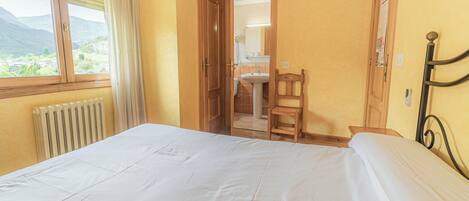 Une chambre à coucher avec un lit, une chaise en bois, une salle de bain avec un lavabo et un miroir, et une vue sur les montagnes par la fenêtre.