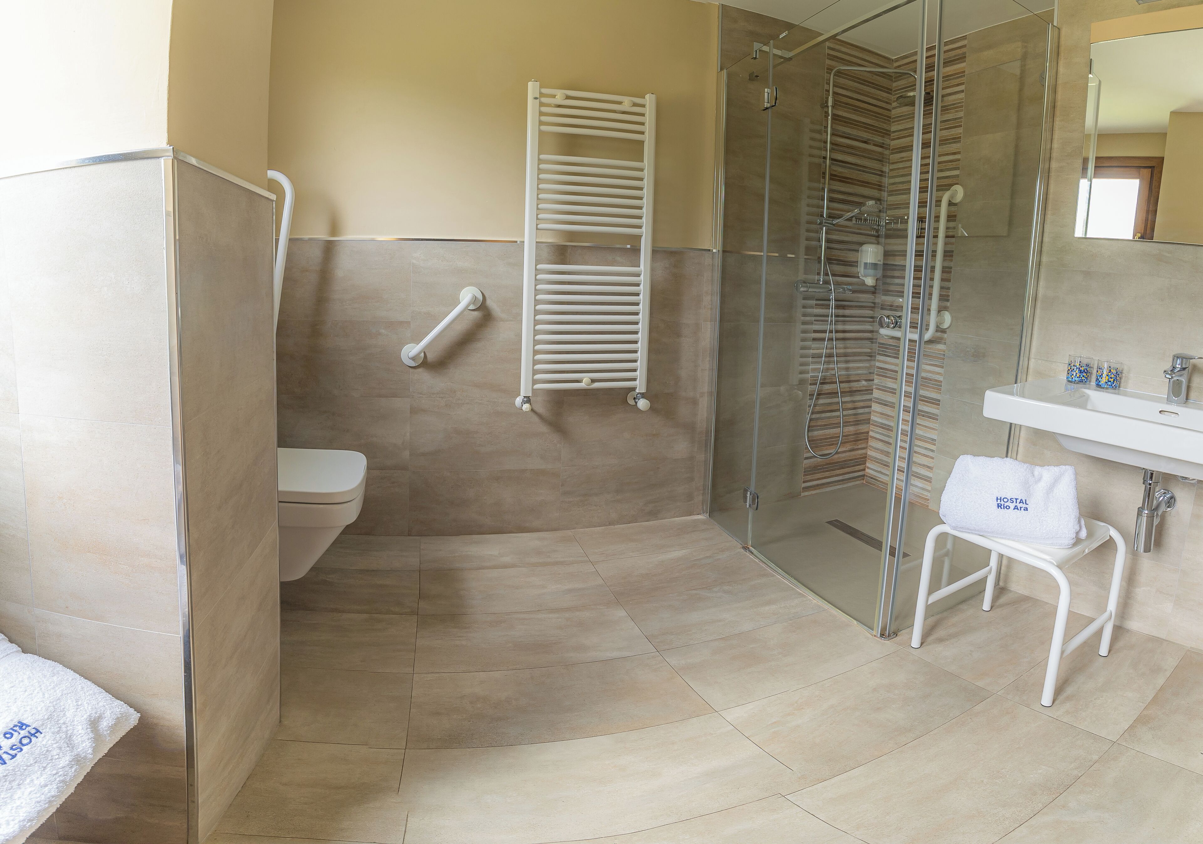 Un baño con cabina de ducha de vidrio, una toalla blanca en el suelo, un taburete blanco con una toalla y un lavabo blanco con espejo.