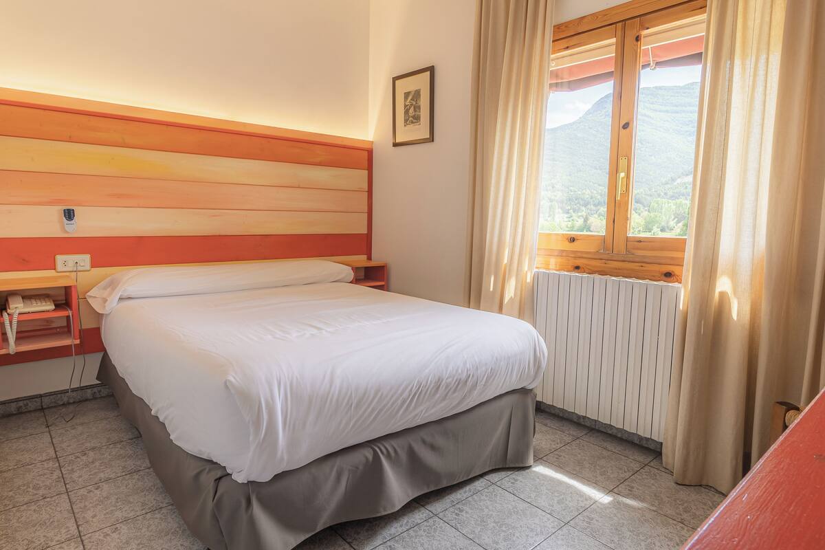 A hotel room with a bed, a wooden headboard, a window with curtains, a radiator, and a telephone on a shelf.