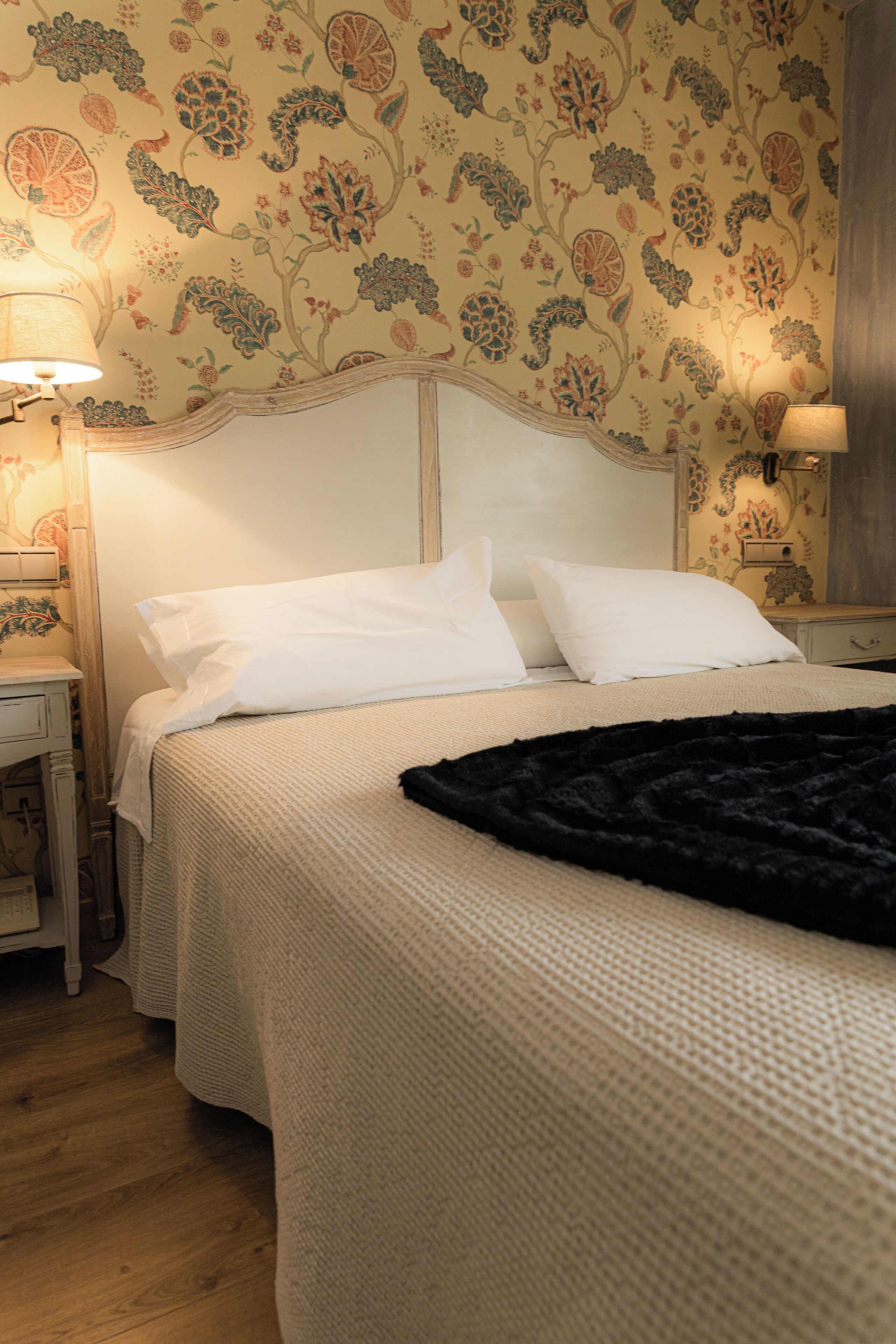 Un lit bien fait, avec du linge de lit blanc et un plaid noir, une table de chevet en bois avec une lampe, et un papier peint à motifs floraux.