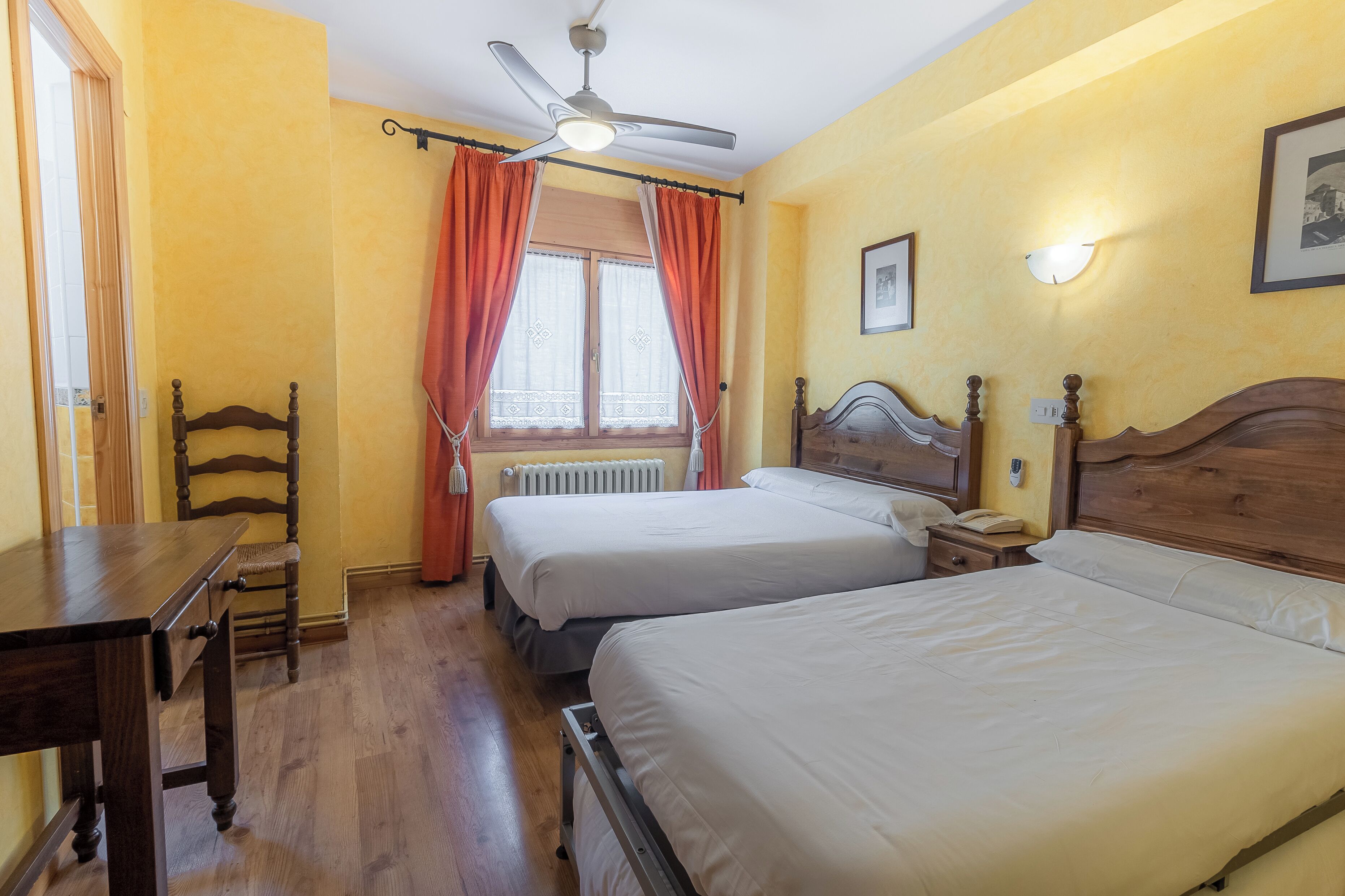 A hotel room with two beds, a wooden table, a chair, a ceiling fan, and a window with curtains.