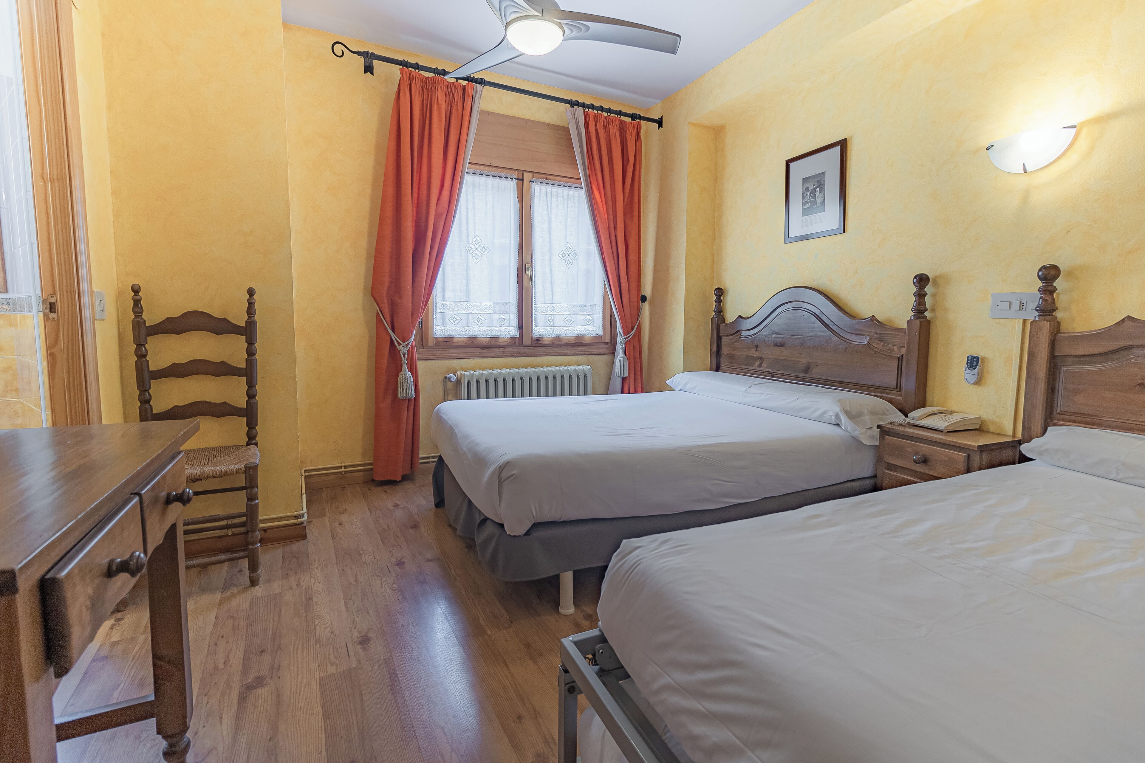 A room with two beds, a wooden chair, a desk, a radiator, and a ceiling fan.