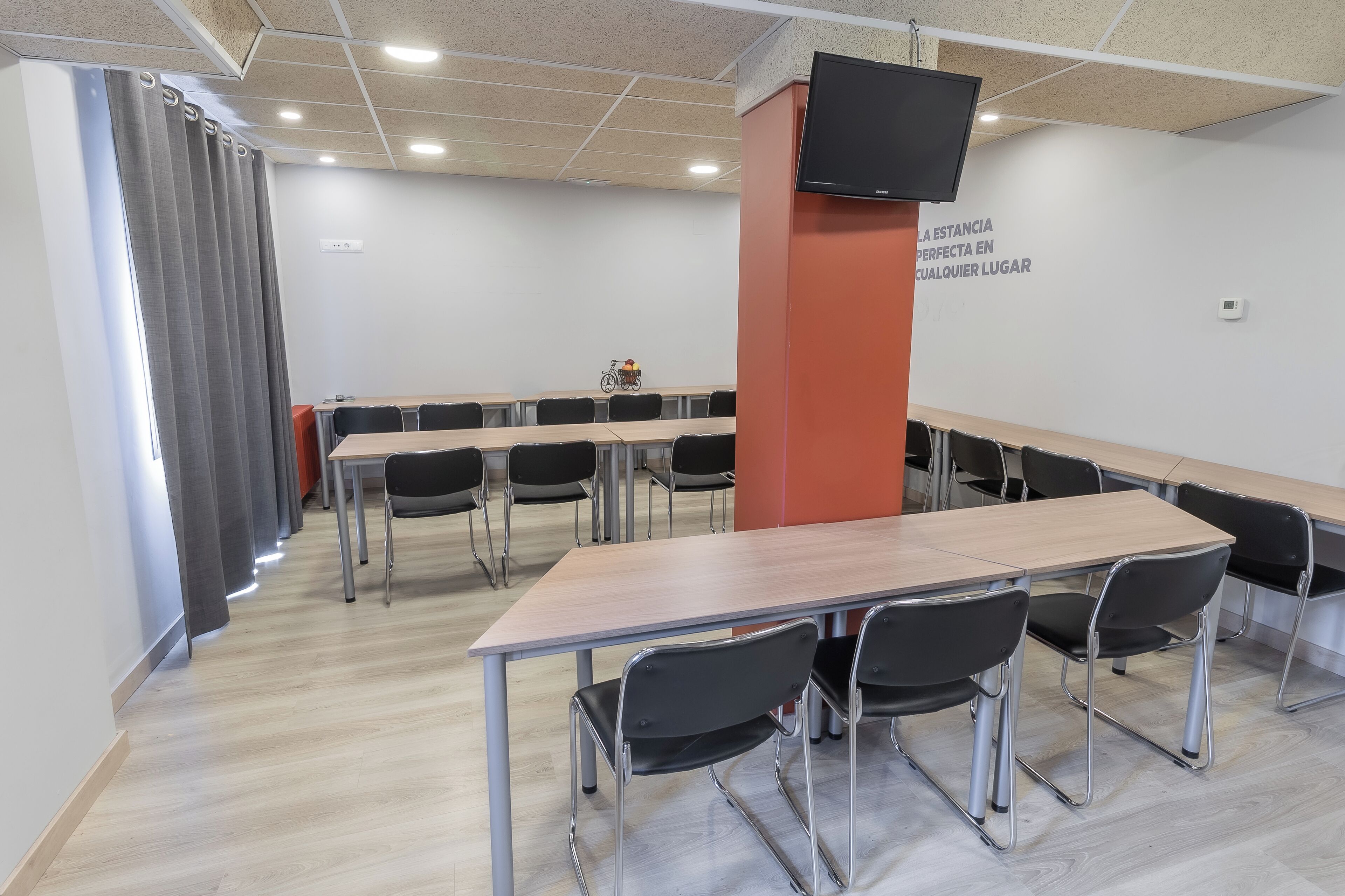 A conference room with a long table, black chairs, a television on the wall, and a window with curtains.