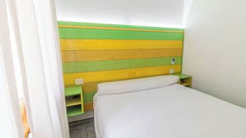 Une petite pièce aux couleurs vives, avec une tête de lit verte et jaune, un lit blanc et une petite table de chevet verte sur laquelle se trouve une lampe.