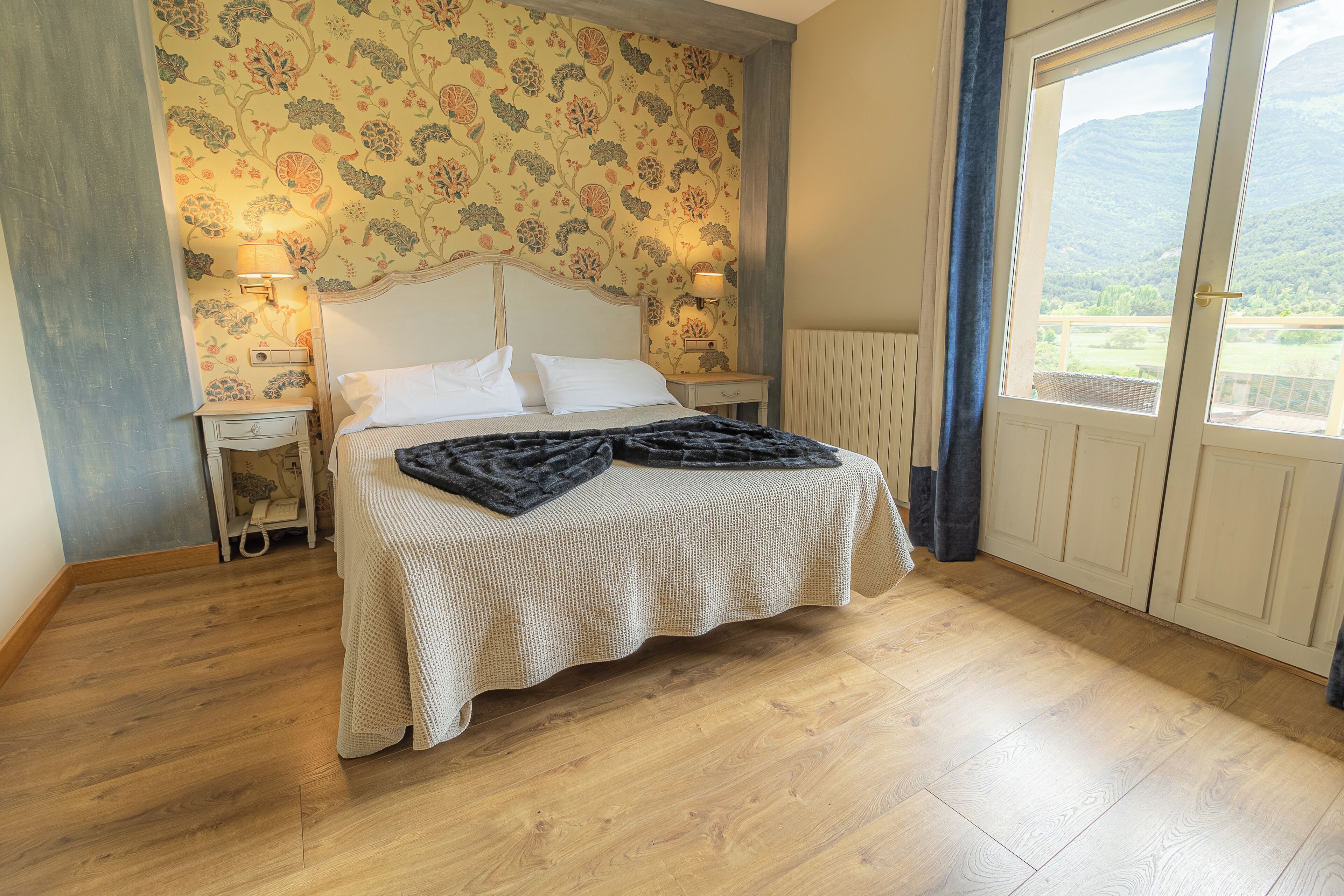 A bedroom with a bed, two bedside tables, a lamp, a window with a view, and a patterned wallpaper.