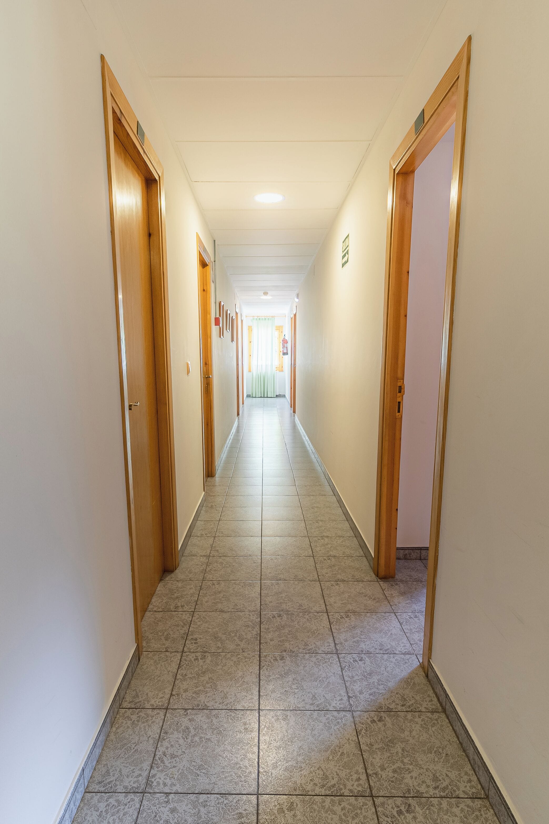 A long, narrow hallway with wooden doors on both sides, tiled flooring, and a ceiling with recessed lighting.