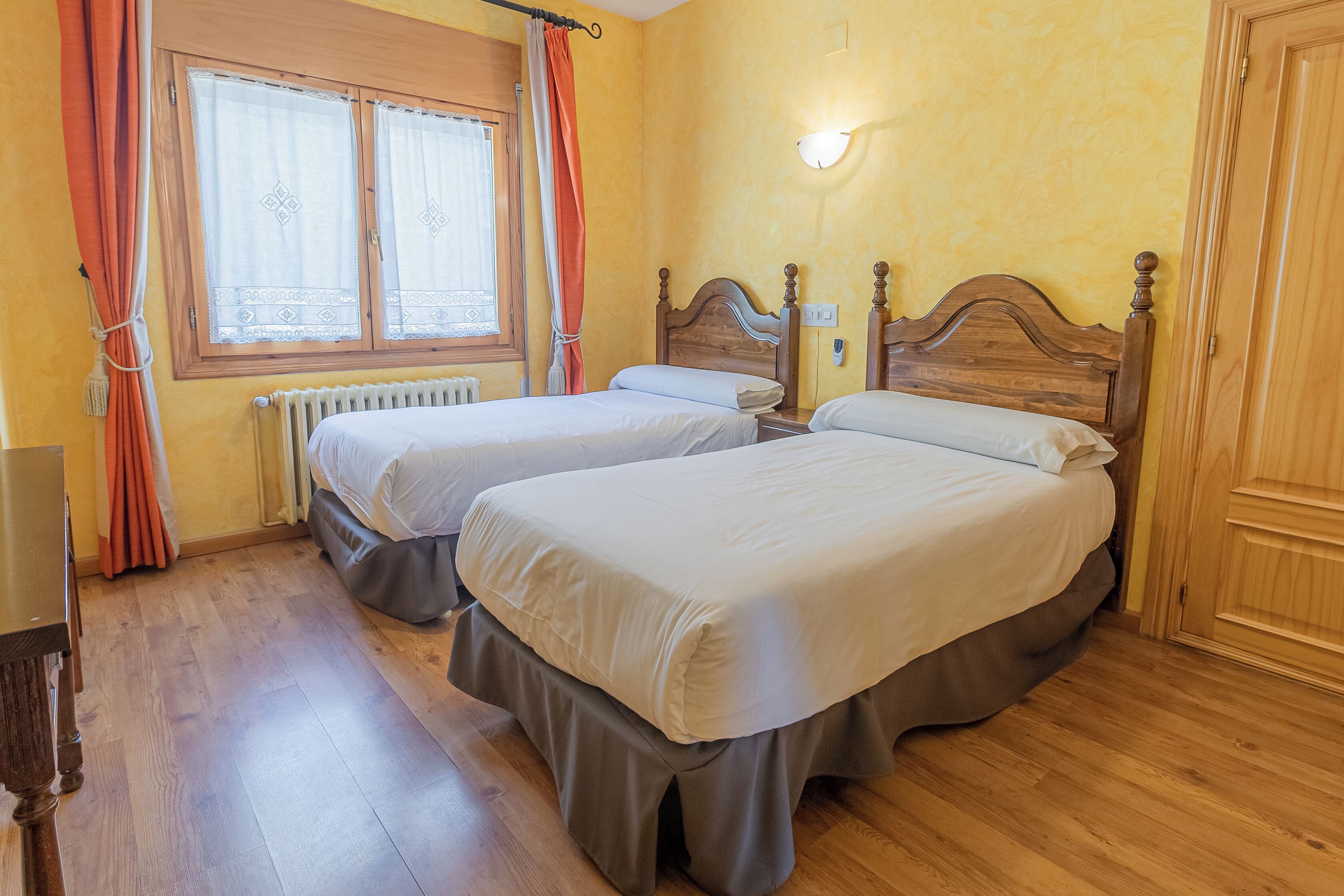 Une chambre avec deux lits, des têtes de lit en bois, une fenêtre avec des rideaux, un radiateur et une porte en bois.