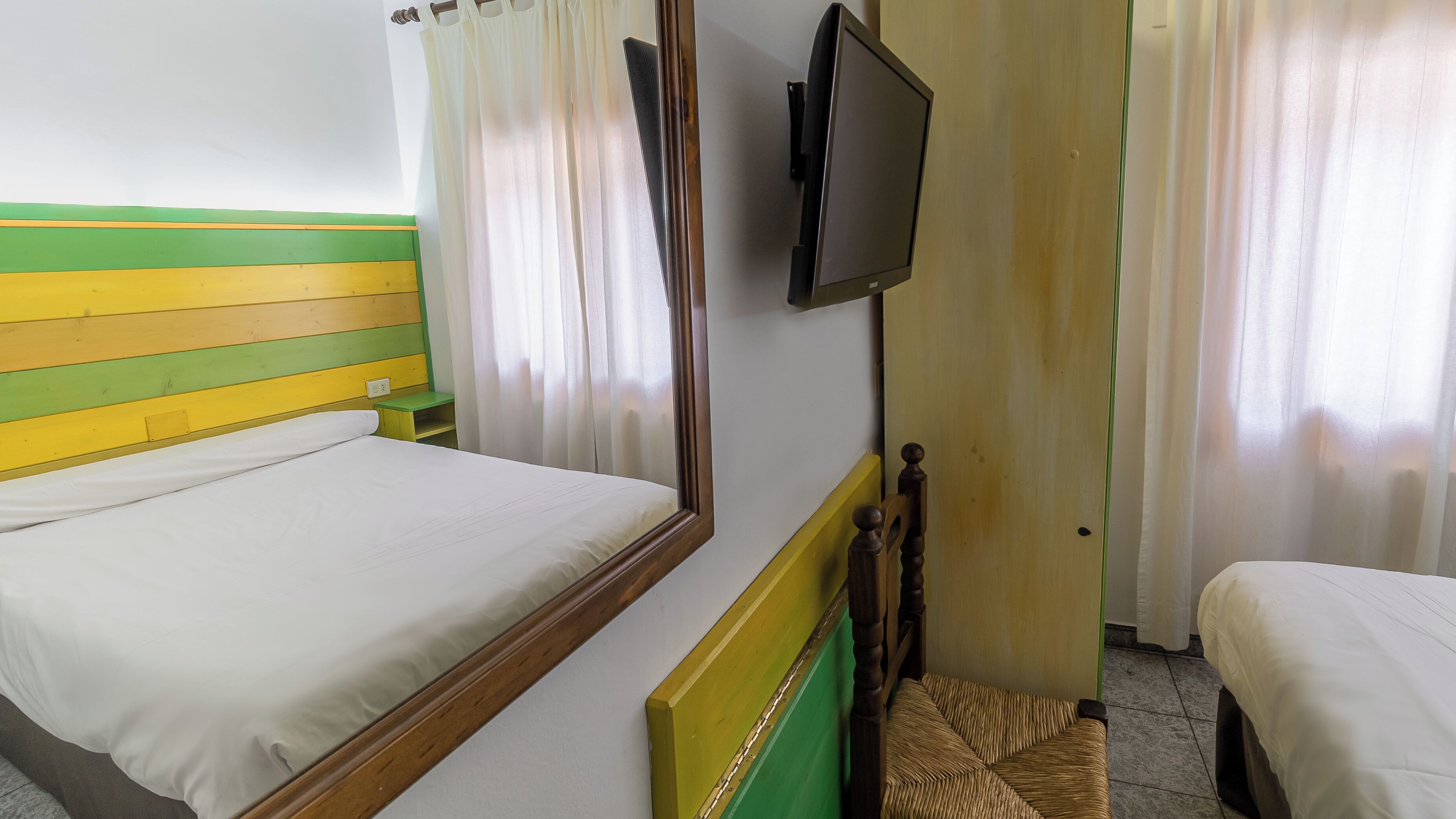 A room with two beds, a television mounted on the wall, and a small wooden bench.