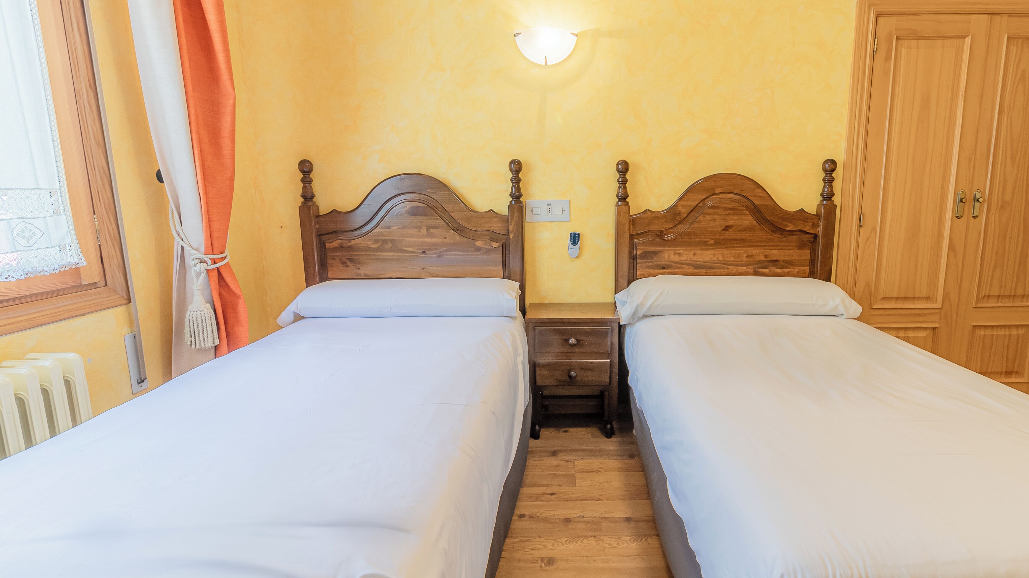 A double room with two wooden beds, a nightstand, a wall-mounted light, and a wooden wardrobe.