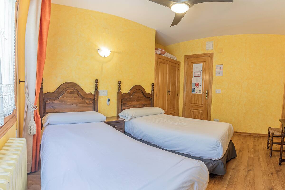 A hotel room with two single beds, wooden headboards, a wooden wardrobe, a desk with a chair, and a ceiling fan.