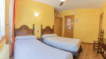 Une chambre d’hôtel avec deux lits simples, des têtes de lit en bois, une armoire en bois, un bureau avec une chaise et un ventilateur de plafond.