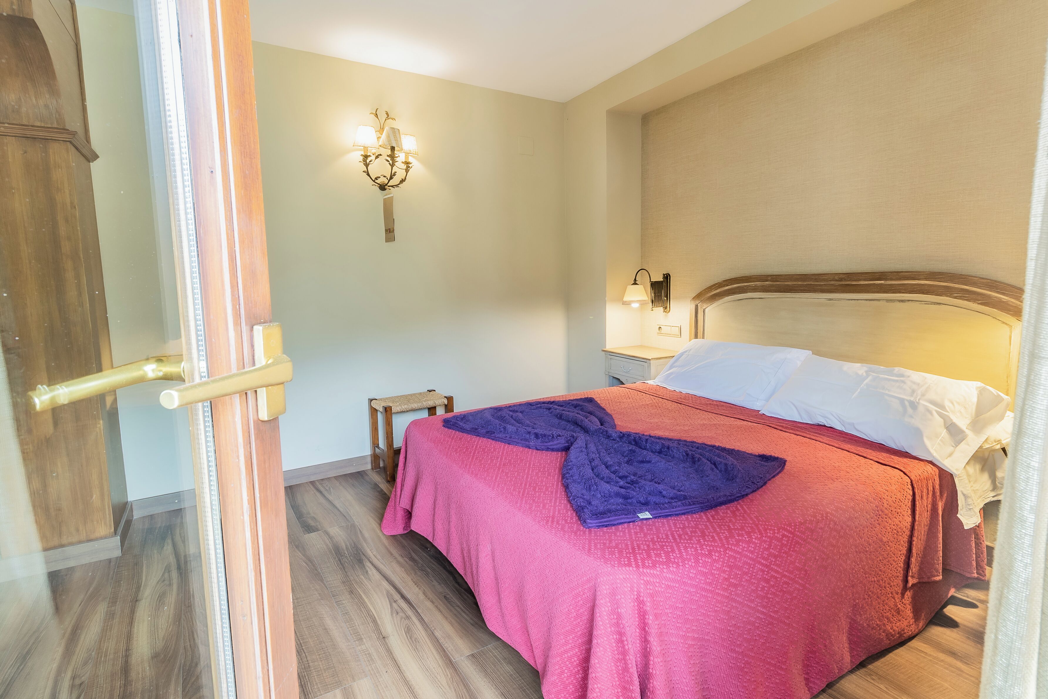 A bedroom with a bed, a nightstand, a wall-mounted lamp, and a door with a brass handle.