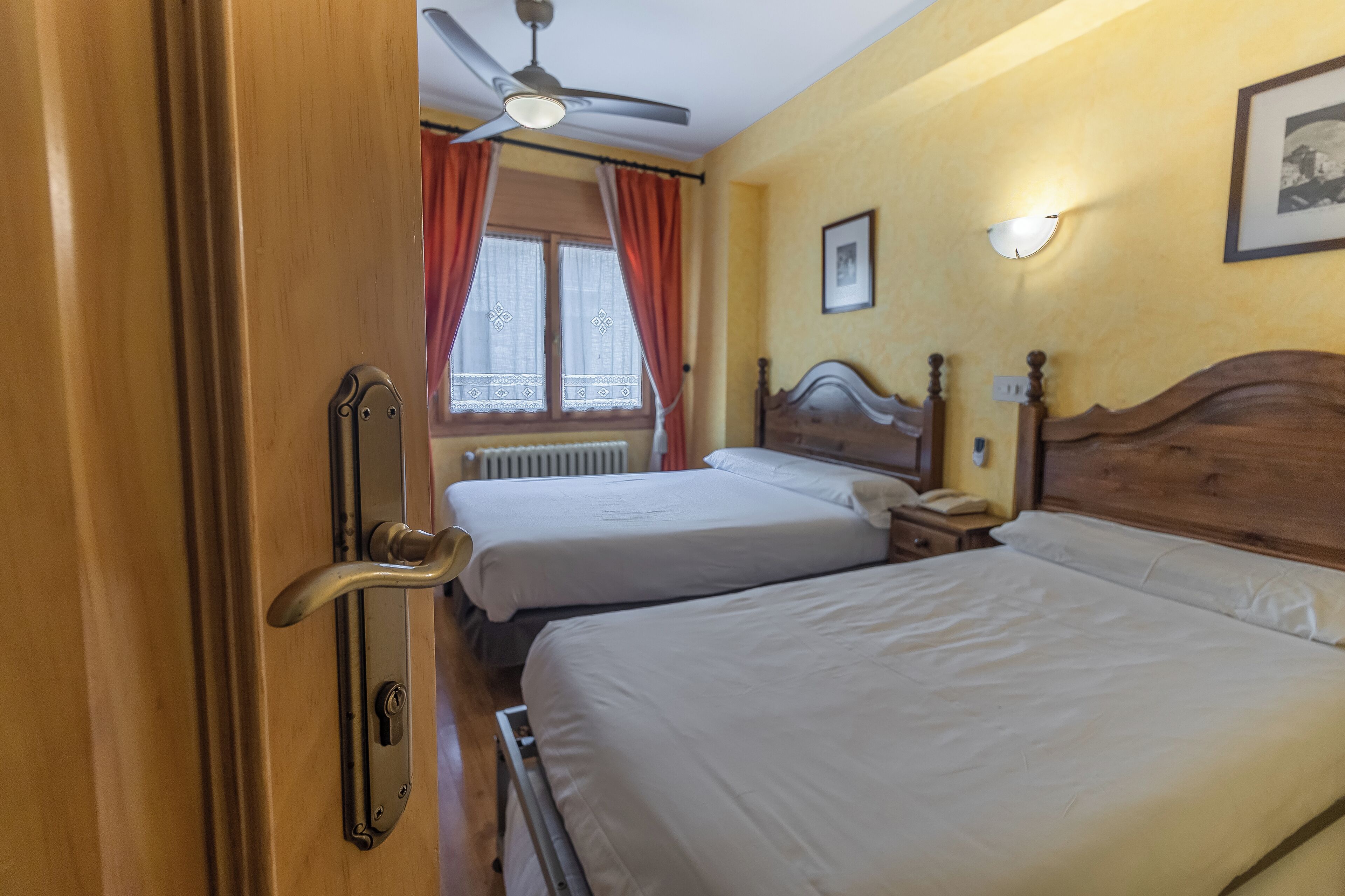 Et hotelværelse med to senge, en ventilator i loftet, et vindue med gardiner, et natbord med lampe samt et billede i ramme på væggen.