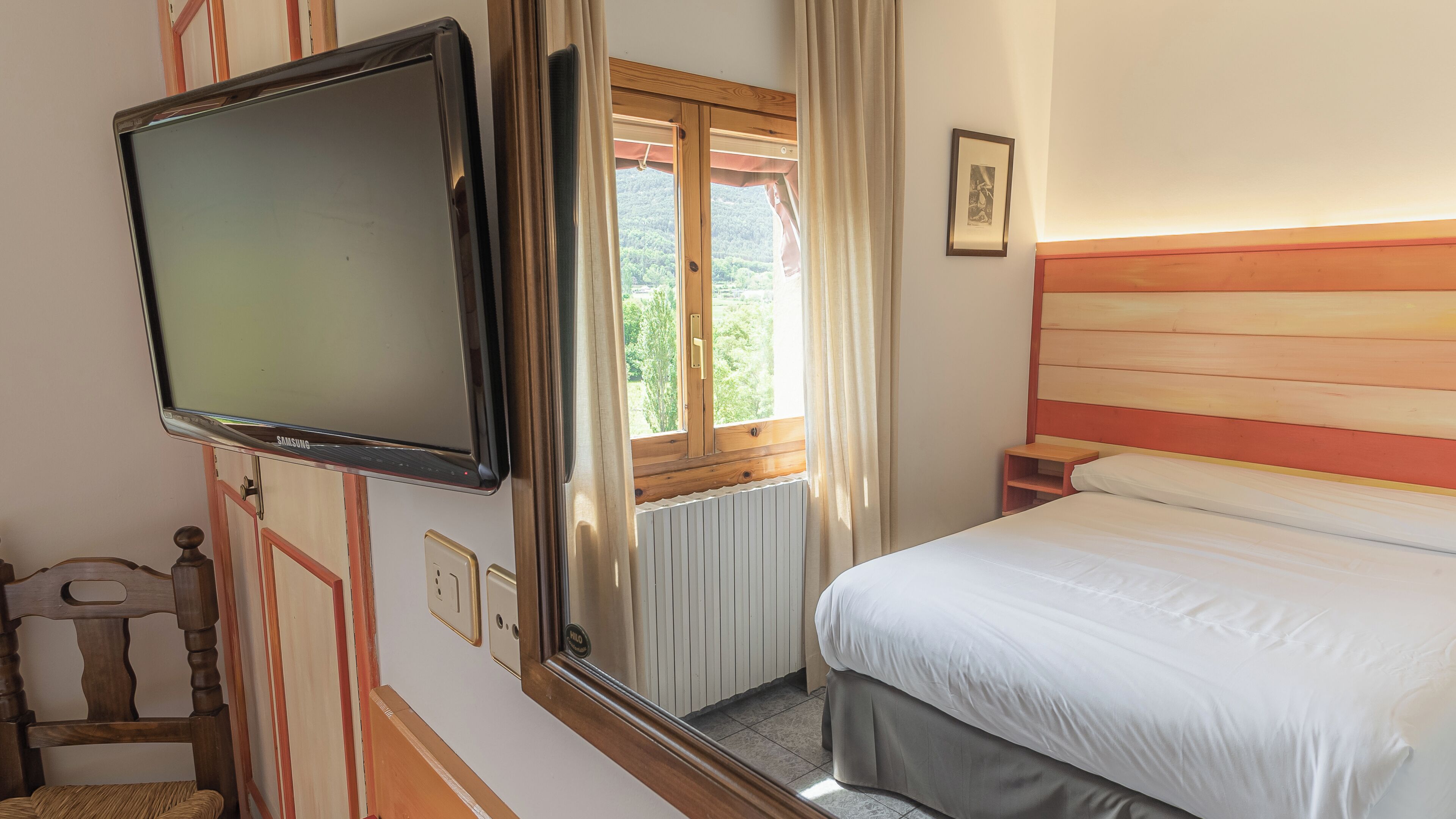 A bedroom with a bed, a TV mounted on the wall, a window with curtains, a bedside table, and a small wooden chair.