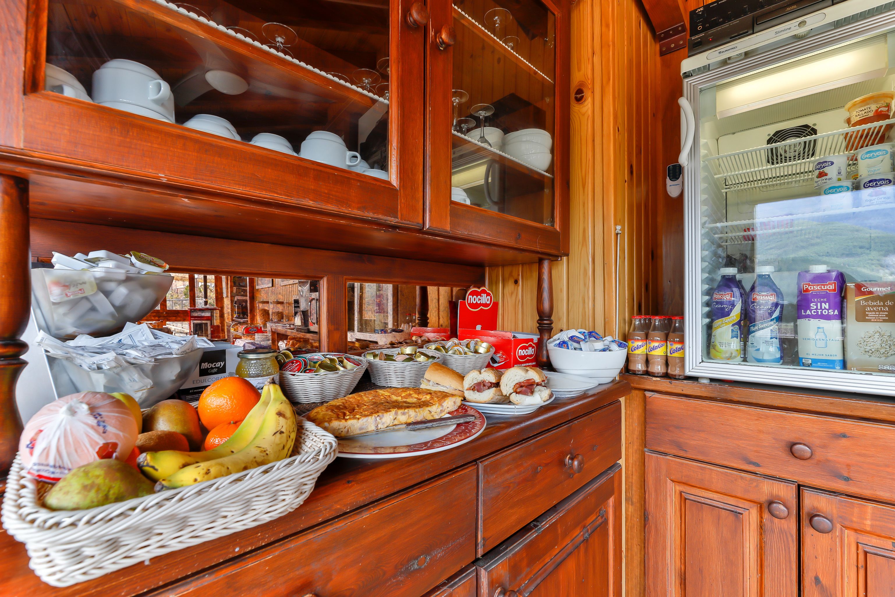 Una cocina con armarios de madera, un refrigerador lleno de bebidas y diversos alimentos en la encimera.