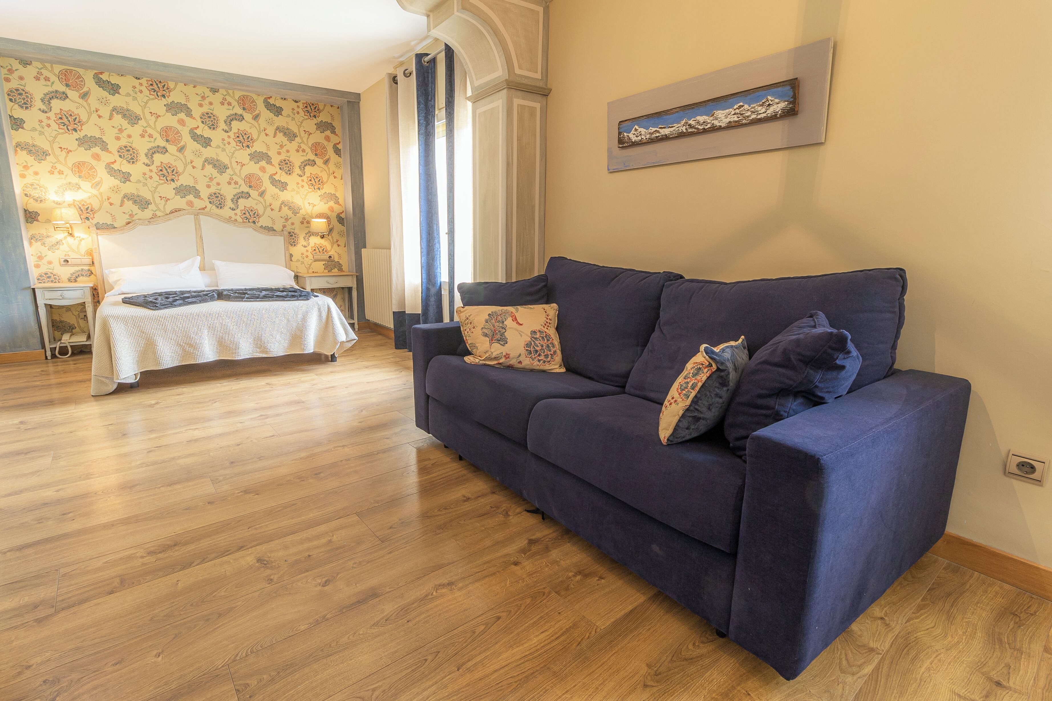Una habitación con cama, sofá azul, suelo de madera y papel tapiz estampado.