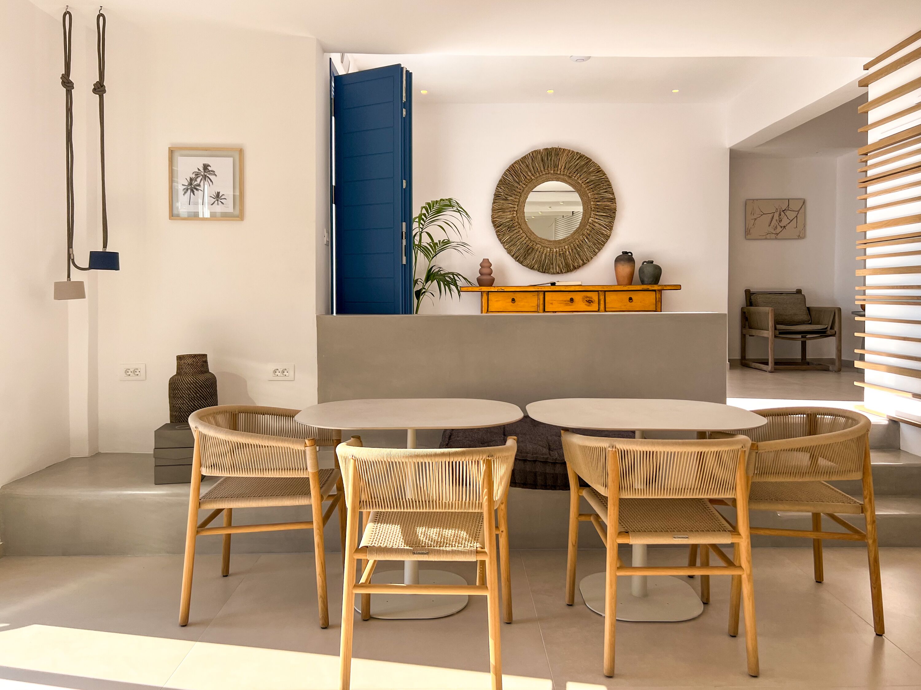 A modern living room with a round dining table, four wicker chairs, a blue door, a woven mirror, and a wooden cabinet.