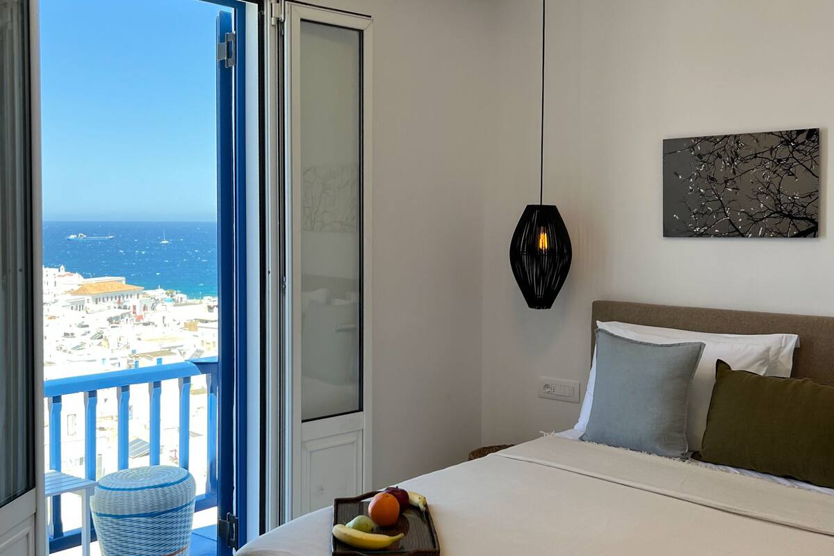 Une chambre avec un grand lit, une vue sur la mer et un balcon agrémenté d’un fauteuil en osier.