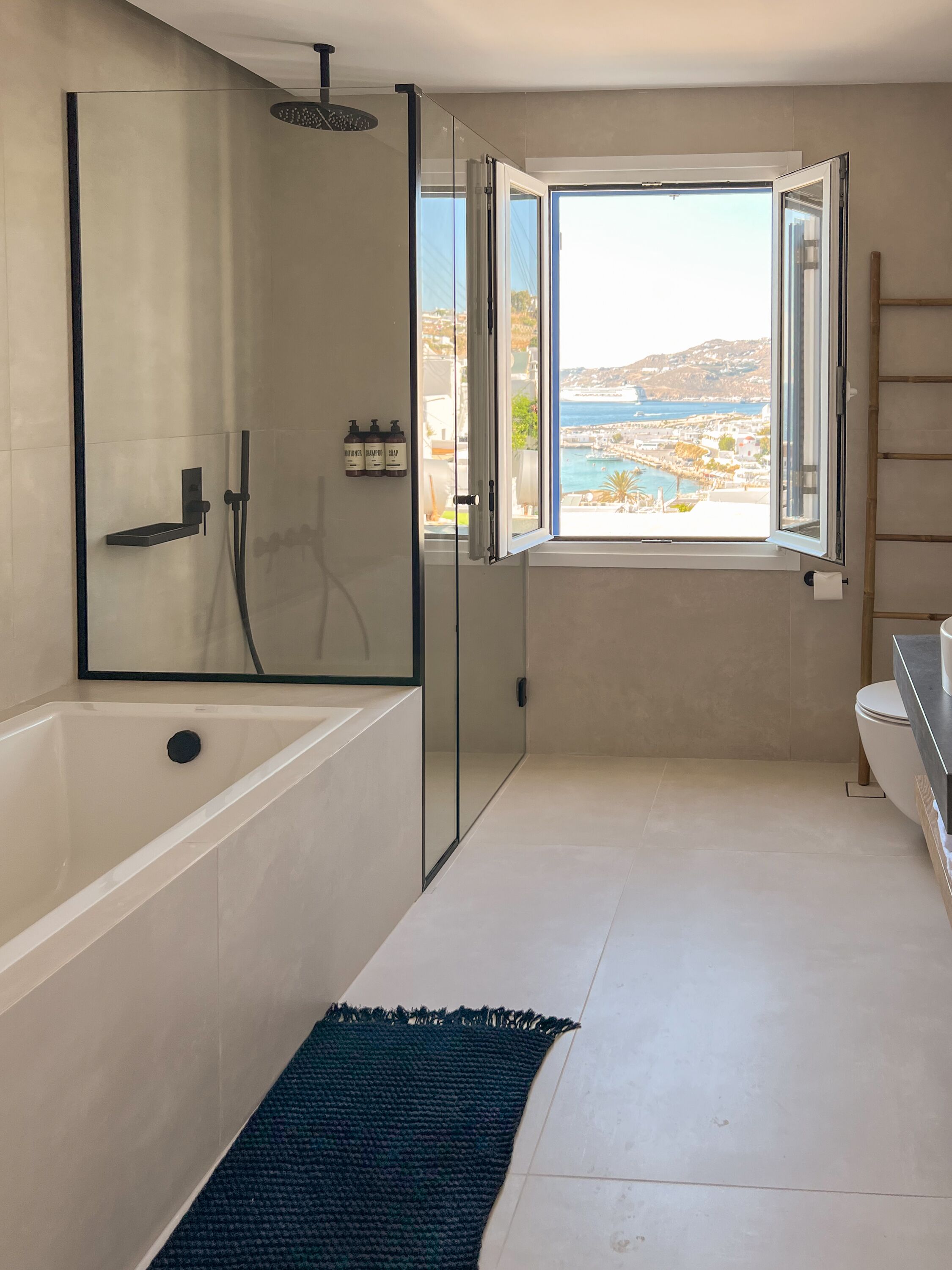 A modern bathroom with a glass-enclosed shower, a large bathtub, a toilet, and a window offering a view of a coastal town.