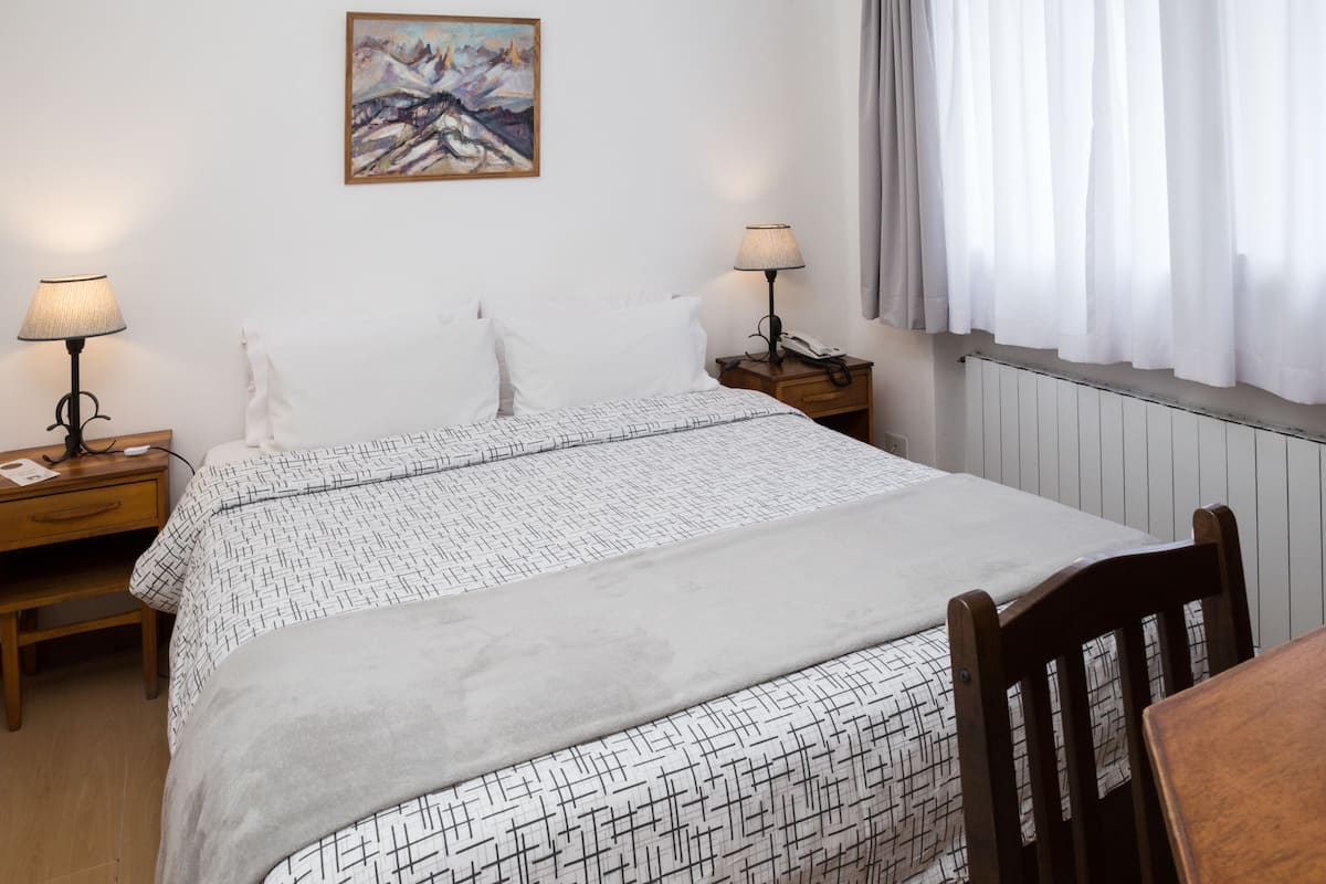 Ein Schlafzimmer mit einem Bett, zwei Nachttischen, einem Sessel, einem Bild an der Wand und einem Fenster mit Vorhängen.