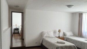 Une chambre d’hôtel avec deux lits simples, un plancher en bois, une petite table avec une lampe et une vue sur le couloir.